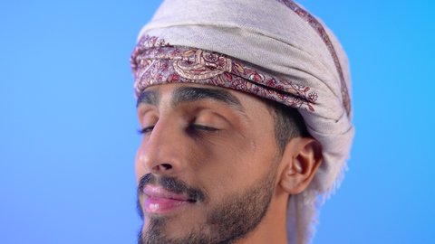 The concept of elegance and interest in outward appearance, looking at the camera with gestures of happiness and pleasure, a portrait of a young Arab Gulf Omani man wearing a dishdasha and turban, standing straight, blue background
