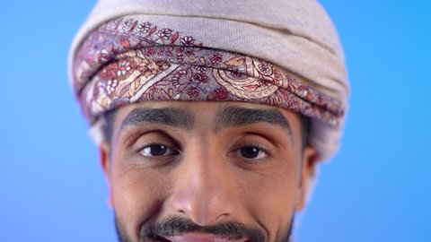 Standing straight and looking at the camera with gestures of happiness and pleasure, the concept of elegance and interest in external appearance, a close-up portrait of a young Arab Gulf Omani man wearing a dishdasha and turban, smiling, blue background