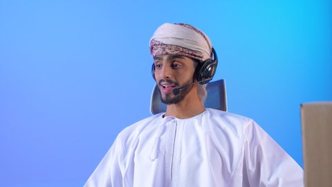 انجاز المهام عن بعد، التواصل مع الأهل والأصدقاء، استخدام التكنولوجيا والأجهزة التقنية الحديثة المتطورة، مفهوم الإصغاء والإستماع, بورتريه لشاب عربي خليجي عماني مبتسم يرتدي الدشداشة و العمامة يجلس على الطاوله امام الحاسب المحمول ويقوم بالتحدث عبر الميكروفون, خلفية زرقاء