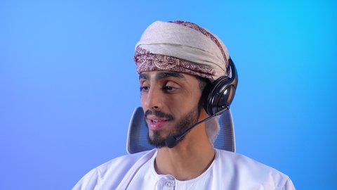 الدردشة مع الأهل والأصدقاء، استخدام تقنيات الهيدفون، مفهوم الإصغاء والإستماع, بورتريه مقرب لشاب عربي خليجي عماني مبتسم يرتدي الدشداشة و العمامة يقوم بالتحدث عبر الميكروفون, خلفية زرقاء