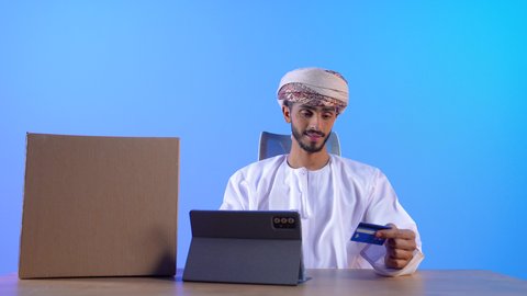 الشراء والدفع عن طريق بطاقات الإئتمان البنكية، إمكانية تسديد الفواتير عبر بطاقة الصراف, بورتريه مقرب لشاب عربي خليجي عماني يرتدي الدشداشة و العمامة يجلس على الطاولة و بجانبة التابلت يمسك بيده البطاقة الالكترونية, خلفية زرقاء