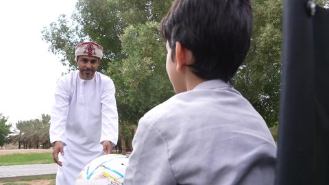 اللعب بكرة القدم بإيماءات الفرح والسرور، ممارسة الأنشطة الترفيهية وقضاء أوقات متعة مع الأبناء,  توفير المستلزمات الخاصة بالمعاقين، نمط حياة ذوي الهمم، ولد عربي خليجي عماني يرتدي الزي العماني التقليدي يجلس على كرسي متحرك في الحديقة برفقة والده