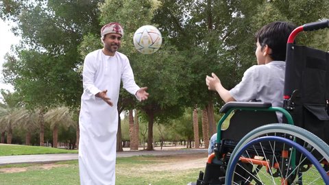 Practicing recreational activities and spending fun times with children, playing football with gestures of joy and happiness, providing supplies for the disabled, the lifestyle of people with disabilities, an Arab Gulf Omani boy wearing traditional Omani clothing sitting in a wheelchair in the park with his father
