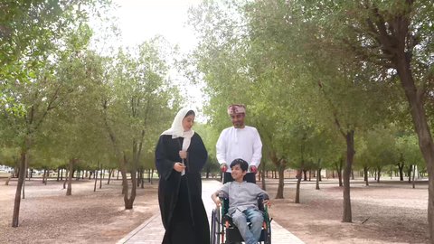 التنزه في احدى الحدائق العامة، الاستمتاع بالأجواء الصيفيه المشمسة، تصوير لعائلة عربية خليجية عمانية تقوم بمساعدة طفل يجلس على الكرسي المتحرك، تأمين ادوات ومعدات تساعد أصحاب الهمم على التنقل والحركة، النظر الى شيء ما