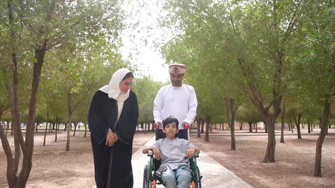 الاستمتاع بالأجواء الصيفيه المشمسة، تصوير لعائلة عربية خليجية عمانية تقوم بمساعدة طفل يجلس على الكرسي المتحرك، تأمين ادوات ومعدات تساعد أصحاب الهمم على التنقل والحركة، النظر الى شيء ما، التنزه في احدى الحدائق العامة