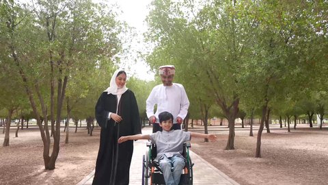تصوير لعائلة عربية خليجية عمانية تقوم بمساعدة طفل يجلس على الكرسي المتحرك، تأمين ادوات ومعدات تساعد أصحاب الهمم على التنقل والحركة، النظر الى شيء ما، التنزه في احدى الحدائق العامة،  الاستمتاع بالأجواء الصيفيه المشمسة