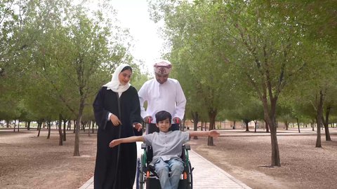 تأمين ادوات ومعدات تساعد أصحاب الهمم على التنقل والحركة، النظر الى شيء ما، التنزه في احدى الحدائق العامة، تصوير لعائلة عربية خليجية عمانية تقوم بمساعدة طفل يجلس على الكرسي المتحرك، الاستمتاع بالأجواء الصيفيه المشمسة