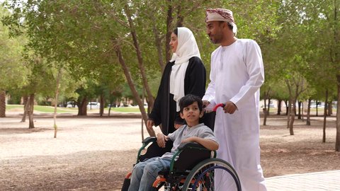 النظر الى شيء ما، التنزه في احدى الحدائق العامة، تصوير لعائلة عربية خليجية عمانية تقوم بمساعدة طفل يجلس على الكرسي المتحرك، الاستمتاع بالأجواء الصيفيه المشمسة، تأمين ادوات ومعدات تساعد أصحاب الهمم على التنقل والحركة