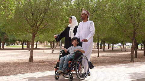 التنزه في احدى الحدائق العامة، تصوير لعائلة عربية خليجية عمانية تقوم بمساعدة طفل يجلس على الكرسي المتحرك، الاستمتاع بالأجواء الصيفيه المشمسة، الاشارة والنظر الى شيء ما، تأمين ادوات ومعدات تساعد أصحاب الهمم على التنقل والحركة