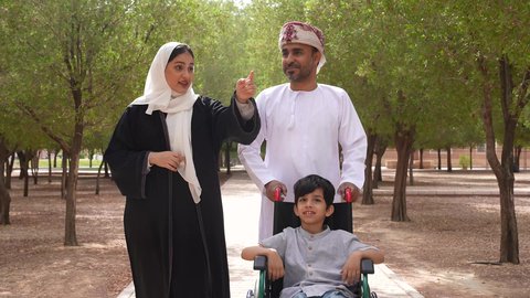 الاستمتاع بالأجواء الصيفيه المشمسة، الاشارة والنظر الى شيء ما، تأمين ادوات ومعدات تساعد أصحاب الهمم على التنقل والحركة، تصوير لعائلة عربية خليجية عمانية تقوم بمساعدة طفل يجلس على الكرسي المتحرك، التنزه في احدى الحدائق العامة