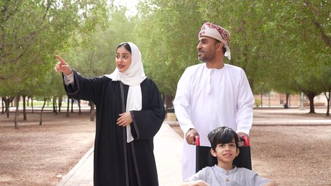 تأمين ادوات ومعدات تساعد أصحاب الهمم على التنقل والحركة، تصوير لعائلة عربية خليجية عمانية تقوم بمساعدة طفل يجلس على الكرسي المتحرك، الاستمتاع بالأجواء الصيفيه المشمسة، التنزه في احدى الحدائق العامة، الاشارة والنظر الى شيء ما