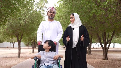 A photo of an Omani Gulf Arab family helping a child in a wheelchair, providing tools and equipment to help people with disabilities move around, enjoying the sunny summer weather, taking a walk in a public park, and looking at something.
