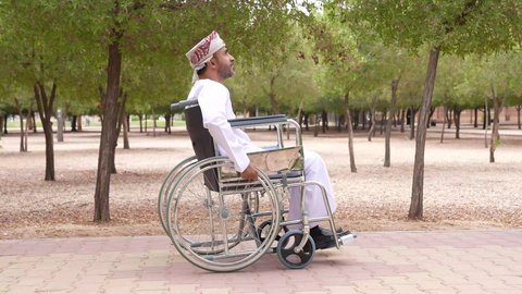التجول والتنزه فى الهواء الطلق، توفير اجهزة ومعدات تساعد بالحركة والتنقل ، العنايه بأصحاب الهمم وتأمين احتياجاتهم الخاصة، تصوير لشاب عربي خليجي عماني مقعد يرتدي الكمه و الدشداشة يجلس على كرسي متحرك
