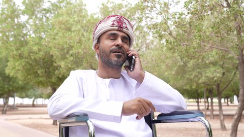 Making phone calls, walking and hiking in the open air, providing devices and equipment that help with movement and mobility, caring for people with disabilities and providing for their special needs, a photo of a disabled young Arab Gulf Omani man wearing a keffiyeh and a dishdasha sitting in a wheelchair