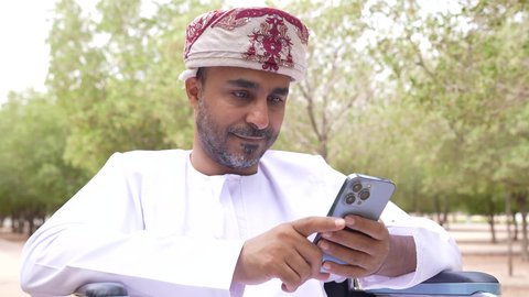 استخدام الهاتف المحمول ، التجول والتنزه فى الهواء الطلق، توفير اجهزة ومعدات تساعد بالحركة والتنقل ، العنايه بأصحاب الهمم وتأمين احتياجاتهم الخاصة، تصوير لشاب عربي خليجي عماني مقعد يرتدي الكمه و الدشداشة يجلس على كرسي متحرك