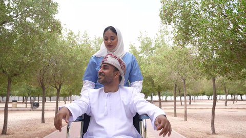 تصوير لممرضة عربية خليجية عمانية محجبة تقوم بمساعدة رجل مريض يجلس على الكرسي المتحرك، تأمين ادوات ومعدات تساعد أصحاب الهمم على التنقل والحركة، الاستمتاع بالأجواء الصيفيه المشمسة، التنزه في احدى الحدائق العامة، النظر الى شيء ما