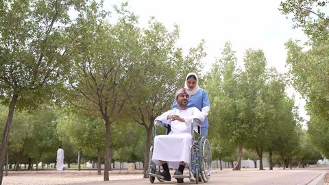 Enjoying the sunny summer weather, walking in a public park, photographing a veiled Gulf Arab Omani nurse helping a sick man sitting in a wheelchair, providing tools and equipment to help people with disabilities move around, looking at something