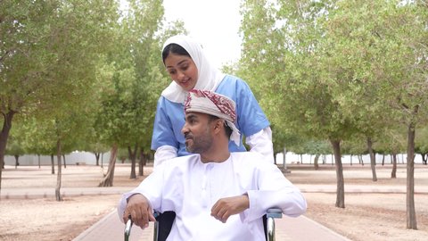 التنزه في احدى الحدائق العامة، الاستمتاع بالأجواء الصيفيه المشمسة، تصوير لممرضة عربية خليجية عمانية محجبة تقوم بمساعدة رجل مريض يجلس على الكرسي المتحرك، تأمين ادوات ومعدات تساعد أصحاب الهمم على التنقل والحركة، النظر الى شيء ما