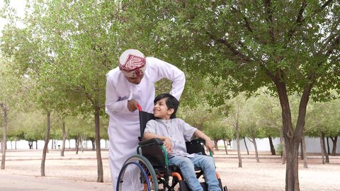 ايماءات السرور والسعادة، الاستمتاع بالأجواء الصيفيه المشمسة، تأمين ادوات ومعدات تساعد أصحاب الهمم على التنقل والحركة، التنزه في احدى الحدائق العامة، النظر الى شيء ما، تصوير لرجل عربي خليجي عماني يقوم بمساعدة طفل يجلس على الكرسي المتحرك