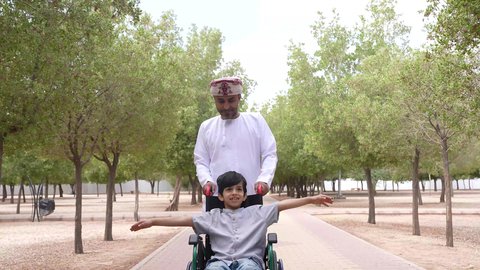 الاستمتاع بالأجواء الصيفيه المشمسة، تأمين ادوات ومعدات تساعد أصحاب الهمم على التنقل والحركة، التنزه في احدى الحدائق العامة، ايماءات السرور والسعادة، النظر الى شيء ما، تصوير لرجل عربي خليجي عماني يقوم بمساعدة طفل يجلس على الكرسي المتحرك
