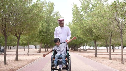 Providing tools and equipment to help people with disabilities move around, taking a walk in a public park, gestures of pleasure and happiness, looking at something, a photo of an Arab Gulf Omani man helping a child sitting in a wheelchair, enjoying the sunny summer weather.