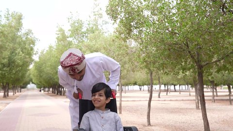 Walking in a public park, gestures of pleasure and happiness, looking at something, a picture of an Arab Gulf Omani man helping a child sitting in a wheelchair, enjoying the sunny summer weather, providing tools and equipment to help people of determination move around.