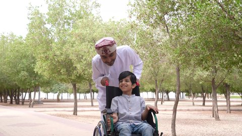 Looking at something, a picture of an Omani Gulf Arab man helping a child sitting in a wheelchair, enjoying the sunny summer weather, providing tools and equipment to help people of determination move around, walking in a public park, gestures of pleasure and happiness