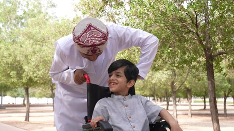 A photo of an Omani Gulf Arab man helping a child sitting in a wheelchair, enjoying the sunny summer weather, providing tools and equipment to help people with disabilities move around, walking in a public park, gestures of pleasure and happiness, looking at something