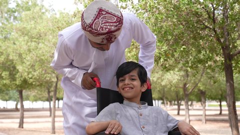 الاستمتاع بالأجواء الصيفيه المشمسة، تأمين ادوات ومعدات تساعد أصحاب الهمم على التنقل والحركة، التنزه في احدى الحدائق العامة، ايماءات السرور والسعادة،  تصوير لرجل عربي خليجي عماني يقوم بمساعدة طفل يجلس على الكرسي المتحرك،  النظر الى شيء ما