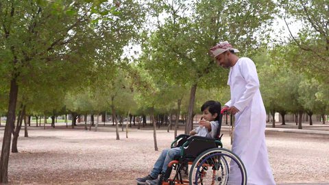 تأمين ادوات ومعدات تساعد أصحاب الهمم على التنقل والحركة، التنزه في احدى الحدائق العامة، ايماءات السرور والسعادة، الاستمتاع بالأجواء الصيفيه المشمسة، تصوير لرجل عربي خليجي عماني يقوم بمساعدة طفل يجلس على الكرسي المتحرك،  النظر الى شيء ما