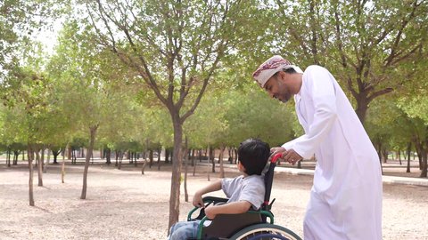 Walking in a public park, gestures of pleasure and happiness, enjoying the sunny summer weather, a picture of an Arab Gulf Omani man helping a child sitting in a wheelchair, providing tools and equipment to help people of determination move around, looking at something