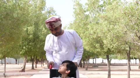 ايماءات السرور والسعادة، التنزه في احدى الحدائق العامة، الاستمتاع بالأجواء الصيفيه المشمسة، تصوير لرجل عربي خليجي عماني يقوم بمساعدة طفل يجلس على الكرسي المتحرك، تأمين ادوات ومعدات تساعد أصحاب الهمم على التنقل والحركة، النظر الى شيء ما