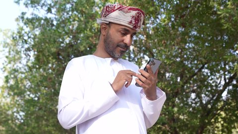 اجراء المكالمات الهاتفية ، الاستمتاع بالأجواء في إحدى المنتزهات، الاستجمام بالطبيعة واستنشاق الهواء الطلق، الخروج إلى أحد الاماكن العامة للترفيه عن النفس، رجل عربي خليجي عماني يرتدي الزي العماني يسير فى احدي حدائق سلطنة عمان