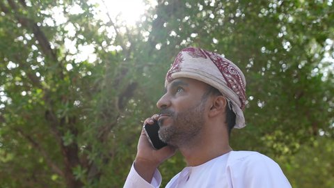Using a mobile phone, enjoying the atmosphere in a park, relaxing in nature and breathing fresh air, going out to a public place to entertain oneself, an Arab Gulf Omani man wearing Omani clothing walking in one of the parks of the Sultanate of Oman