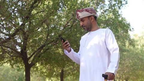 التجول والتنزه فى الهواء الطلق ،اجراء المكالمات الهاتفية ،الاستمتاع بالأجواء في إحدى المنتزهات ،  رجل عربي خليجي عماني يستند على عكاز ، يرتدي الزي العماني يسير فى احدي حدائق سلطنة عمان، الخروج إلى أحد الاماكن العامة للترفيه عن النفس