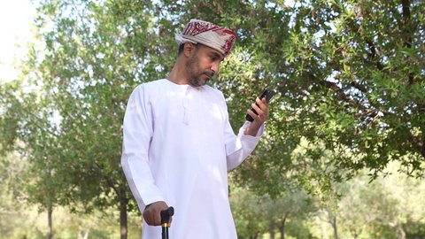 اجراء المكالمات الهاتفية ،  التجول والتنزه فى الهواء الطلق ،الاستمتاع بالأجواء في إحدى المنتزهات ،  رجل عربي خليجي عماني يستند على عكاز ، يرتدي الزي العماني يسير فى احدي حدائق سلطنة عمان، الخروج إلى أحد الاماكن العامة للترفيه عن النفس