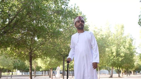Walking and strolling in the open air, enjoying the atmosphere in a park, the concept of recreation in nature, an Arab Gulf Omani man leaning on a crutch, wearing Omani clothing walking in one of the parks of the Sultanate of Oman, going out to a public place to entertain himself