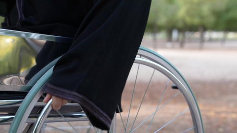 A sense of belonging, appreciation, and respect for people with disabilities in the Sultanate of Oman. Caring for people of determination and ensuring their special needs are met. Equipment and devices are provided to aid mobility. A photograph of a sick Omani Arab Gulf woman wearing a hijab and abaya sitting in a wheelchair, enjoying the atmosphere in a park.