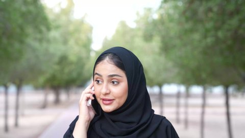 Integrating modern technology into daily life, making phone calls, a young Saudi woman going out to a public place for entertainment, a photograph from behind of an Omani Gulf Arab woman wearing Omani dress walking in one of the parks of the Sultanate of Oman, relaxing in nature and breathing fresh air.