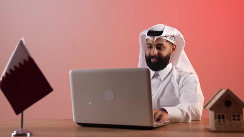 إقامة الاجتماعات أونلاين عبر مكالمات الفيديو، استخدام اللابتوب في العمل، الرد والإجابة على الأسئلة والاستفسارات والمكالمات، بورتريه لرجل عربي خليجي قطري يرتدي الثوب التقليدي والغترة يجلس خلف المكتب أمامه الحاسوب المحمول وعلم قطر ومجسم منزل، خلفية ملونة