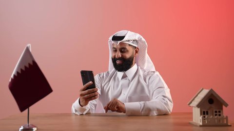 بيع وشراء العقارات، مفهوم الاسكان والتمليك، تصفح مواقع التواصل الاجتماعي عبر الانترنت، استخدام الهاتف المحمول في العمل، بورتريه لرجل عربي خليجي قطري يرتدي الثوب التقليدي والغترة يجلس خلف المكتب أمامه علم قطرومجسم منزل خشبي، خلفية ملونة