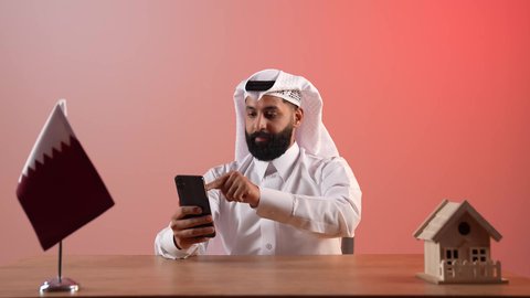 بيع وشراء العقارات، مفهوم الاسكان والتمليك، تصفح مواقع التواصل الاجتماعي عبر الانترنت، استخدام الهاتف المحمول في العمل، بورتريه لرجل عربي خليجي قطري يرتدي الثوب التقليدي والغترة يجلس خلف المكتب أمامه علم قطرومجسم منزل خشبي، خلفية ملونة