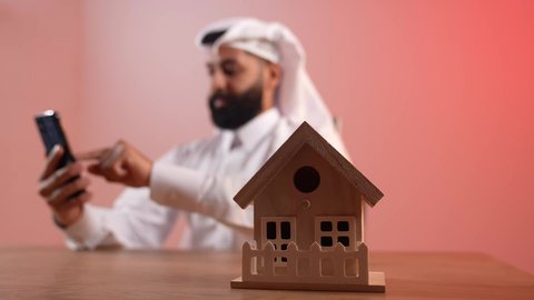 مفهوم الاسكان والتمليك، بيع وشراء العقارات، مجسم منزل خشبي، شاب سعودي يتصفح مواقع التواصل الاجتماعي عبر الانترنت يإيماءات السرور، بورتريه لرجل عربي خليجي قطري يرتدي الثوب التقليدي والغترة يجلس خلف المكتب يقوم باستخدام الهاتف المحمول في العمل، خلفية ملونة