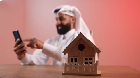 مفهوم الاسكان والتمليك، بيع وشراء العقارات، مجسم منزل خشبي، شاب سعودي يتصفح مواقع التواصل الاجتماعي عبر الانترنت يإيماءات السرور، بورتريه لرجل عربي خليجي قطري يرتدي الثوب التقليدي والغترة يجلس خلف المكتب يقوم باستخدام الهاتف المحمول في العمل، خلفية ملونة