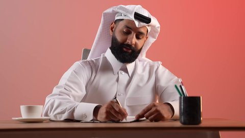 Business and project management concept, portrait of a Saudi Arabian Gulf businessman wearing traditional thobe and ghutra sitting in front of his desk, a young Qatari man taking notes, completing office work tasks, colorful background