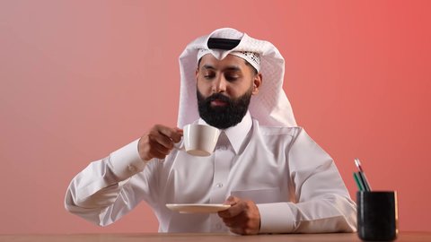 الاسترخاء وتناول مشروبات دافئة، شاب سعودي يقوم بالاستمتاع بكوب من القهوة الساخنة، بورتريه لرجل عربي خليجي قطري يرتدي الثوب التقليدي والغترة يجلس خلف طاولة المكتب ويمسك بيده فنجان من مشروبه المفضل، خلفية ملونة