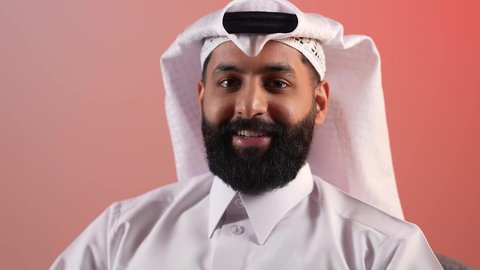 الاهتمام بالأناقة والمظهر الخارجي، بورتريه لرجل عربي خليجي قطري  يرتدي الثوب التقليدي والغترة يجلس على كرسي مريح، شاب سعودي ينظر إلى الكاميرا بإيماءات السرور والثقة في النفس، مفهوم الوسامة والرجولة والقوة، خلفية ملونة