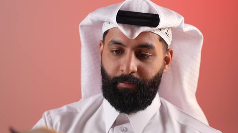 زيادة الذخيرة الفكرية، شاب سعودي يقوم بقراءة الكتب والقصص والروايات، تنمية المهارات وتوسيع المعارف والأفكار، بورتريه مقرب لرجل عربي خليجي قطري يرتدي الثوب التقليدي والغترة يجلس خلف طاولة المكتب ويمسك بيديه كتاب، قضاء أوقات الفراغ بشكل مفيد، خلفية ملونة
