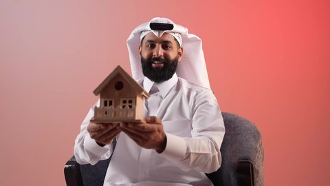 مفهوم الاسكان والتمليك، امتلاك سكن حديث والاستقرار العائلي، شاب قطري ينظر إلى الكاميرا بايماءات السعادة بشراء عقار جديد, بورتريه لرجل عربي خليجي سعودي يرتدي الثوب التقليدي والشماغ يجلس على كرسي مريح يحمل بيده مجسم بيت جديد، خلفية ملونة
