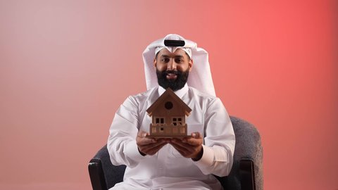 مفهوم الاسكان والتمليك، امتلاك سكن حديث والاستقرار العائلي، شاب قطري ينظر إلى الكاميرا بايماءات السعادة بشراء عقار جديد, بورتريه لرجل عربي خليجي سعودي يرتدي الثوب التقليدي والشماغ يجلس على كرسي مريح يحمل بيده مجسم بيت جديد، خلفية ملونة